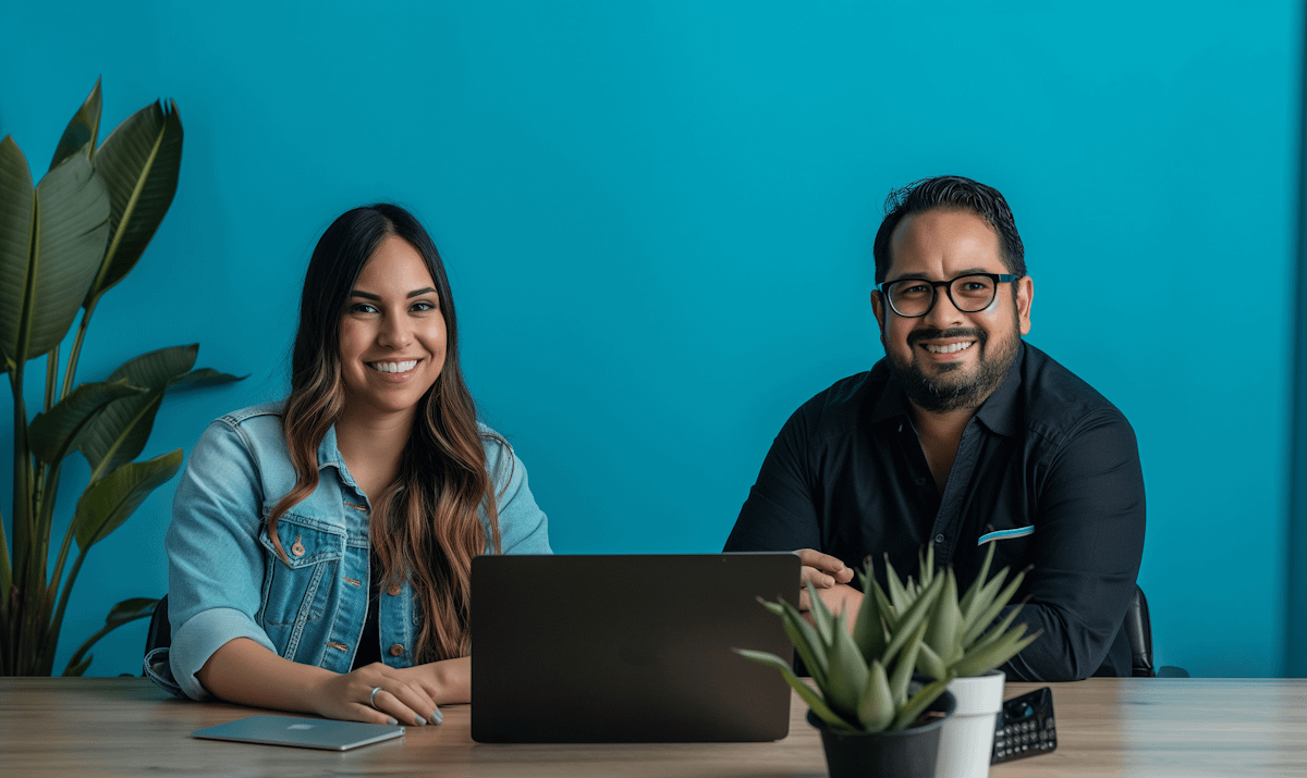 Smiling man and woman at a desk with laptops, vibrant blue background, and a potted plant