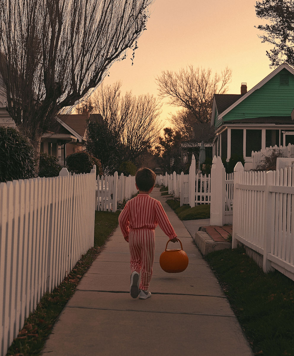 Child in striped pajamas with pumpkin bucket walks down a suburban sidewalk at sunset, surrounded by white picket fences and bare trees