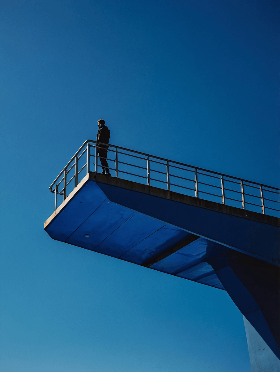 A person stands on a blue diving platform against a vivid blue sky